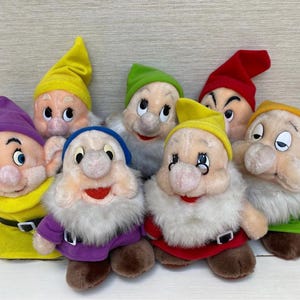 Könnte beinhalten: Eine Sammlung von sieben Zwergen-Plüschpuppen, jede mit einer einzigartigen Hut-Farbe: lila, gelb, grün, rot und blau. Die Puppen haben weiße Bärte, rosige Wangen und tragen bunte Outfits. Ein Etikett ist auf einer der Puppen sichtbar.