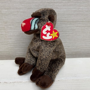 May include: A brown plush toy baboon with a red nose and a tag. The baboon has a speckled pattern on its body and dark brown feet and hands. The tag is red with white text and a yellow star.