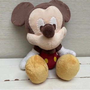 May include: A plush Mickey Mouse toy. It has a brown head and ears, a tan face, and a red body with yellow buttons. The toy has white gloves and tan feet. It is sitting on a white wooden surface.