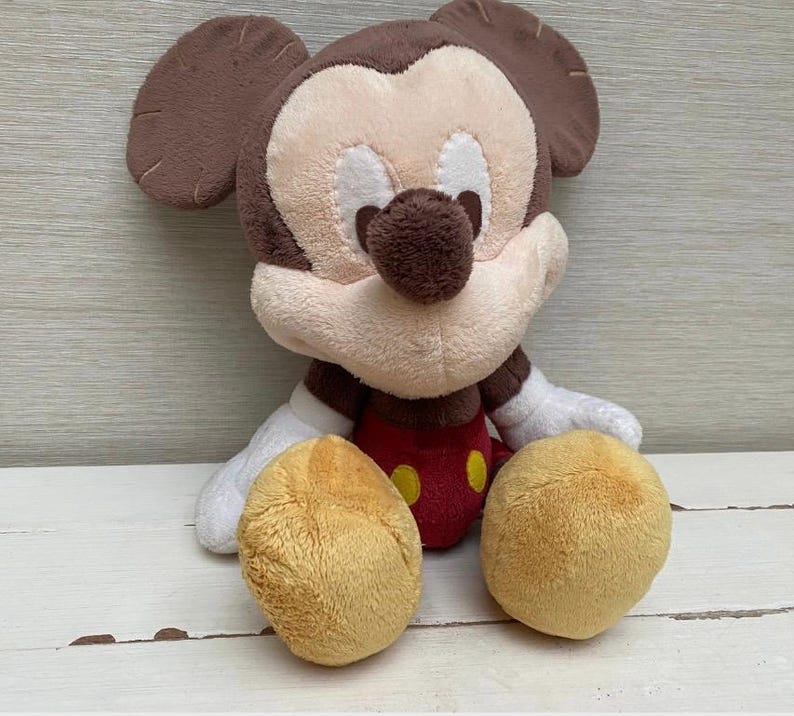 May include: A plush Mickey Mouse toy. It has a brown head and ears, a tan face, and a red body with yellow buttons. The toy has white gloves and tan feet. It is sitting on a white wooden surface.