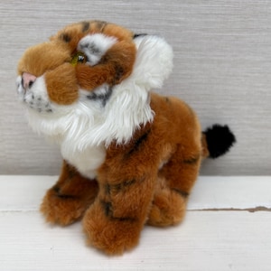 Op de afbeelding: Een pluche tijgerknuffel met oranje en bruine vacht met zwarte strepen, een witte borst en nekpluis, een roze neus en gele ogen. Het knuffeldier zit in een zittende houding met een kleine staart met een zwart uiteinde aan de achterkant.