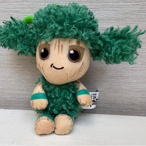 May include: A plush toy of the character Groot from Guardians of the Galaxy. The toy has a tan face with large black eyes and a small smile. It features green, fluffy hair and accents, and is wearing green armbands.