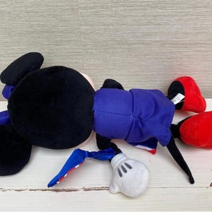 Puede incluir: Un mu&ntilde;eco de peluche de Mickey Mouse, tumbado de lado. El mu&ntilde;eco tiene la cabeza y las orejas negras, un traje azul, zapatos rojos y guantes blancos. El mu&ntilde;eco est&aacute; sobre una superficie de madera blanca.