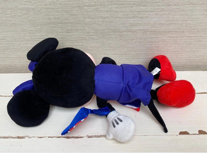 Puede incluir: Un mu&ntilde;eco de peluche de Mickey Mouse, tumbado de lado. El mu&ntilde;eco tiene la cabeza y las orejas negras, un traje azul, zapatos rojos y guantes blancos. El mu&ntilde;eco est&aacute; sobre una superficie de madera blanca.