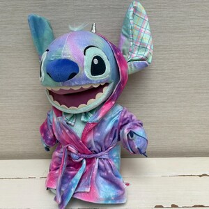 May include: A plush Stitch doll wearing a colorful robe with a matching hood and bow tie. The doll has large ears, a blue nose, and a wide smile. The robe is a mix of pink, purple, and blue, with a tie at the waist.