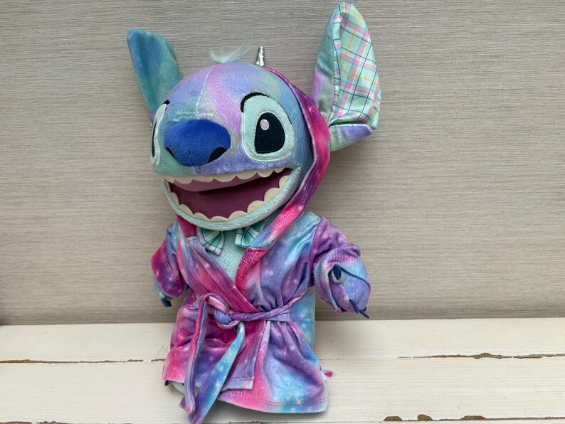 May include: A plush Stitch doll wearing a colorful robe with a matching hood and bow tie. The doll has large ears, a blue nose, and a wide smile. The robe is a mix of pink, purple, and blue, with a tie at the waist.