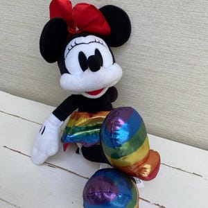 May include: A Minnie Mouse soft toy featuring a red bow, black ears, and a rainbow-striped skirt and shoes. The toy has white gloves and a white face. It is positioned on a white wooden surface.