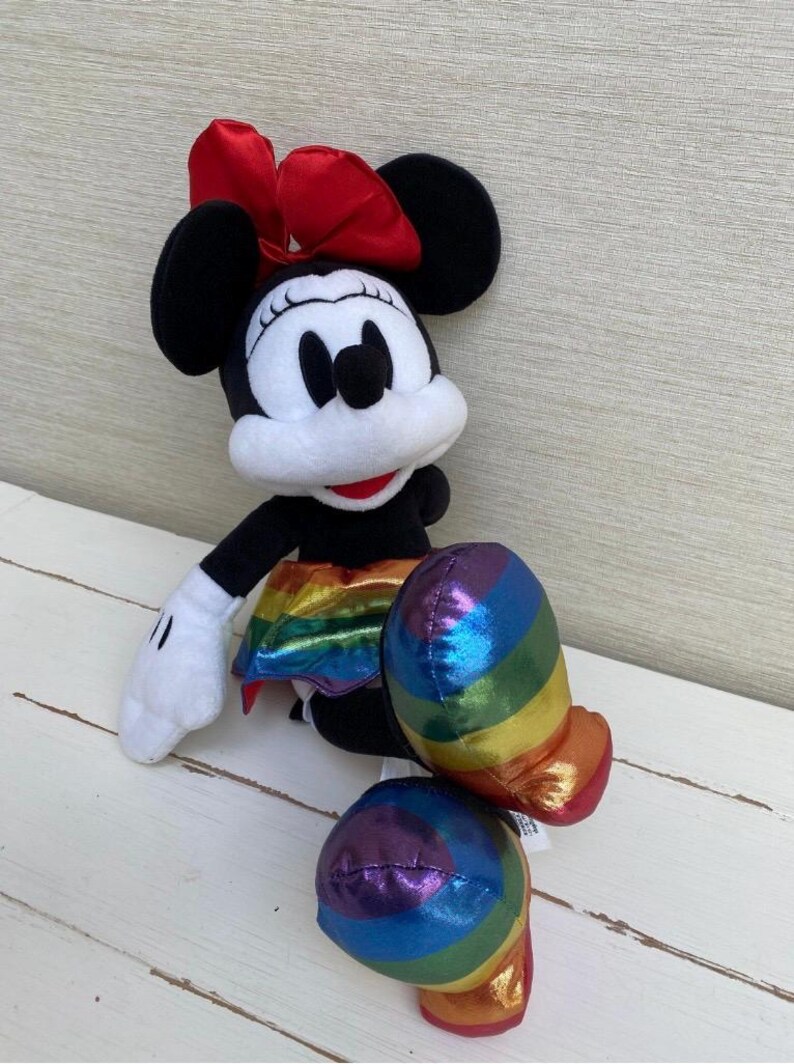 May include: A Minnie Mouse soft toy featuring a red bow, black ears, and a rainbow-striped skirt and shoes. The toy has white gloves and a white face. It is positioned on a white wooden surface.