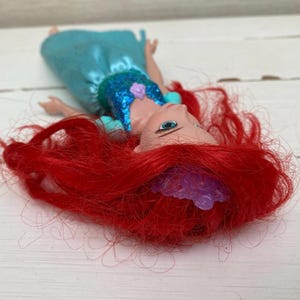 Op de afbeelding: Een pop met felrood haar en een turquoise jurk. De pop heeft een blauwe top met pailletten en een paarse tiara. Ze ligt op een wit houten oppervlak.
