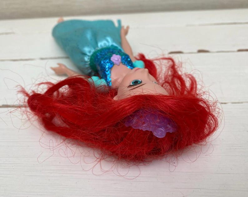 Op de afbeelding: Een pop met felrood haar en een turquoise jurk. De pop heeft een blauwe top met pailletten en een paarse tiara. Ze ligt op een wit houten oppervlak.