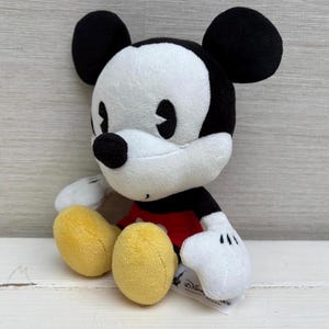 Op de afbeelding: Een pluche Mickey Mouse-speelgoed met zwarte oren, een zwarte neus en een wit gezicht. Het speelgoed heeft een rood shirt, witte handschoenen en gele schoenen. Het speelgoed staat op een wit oppervlak.