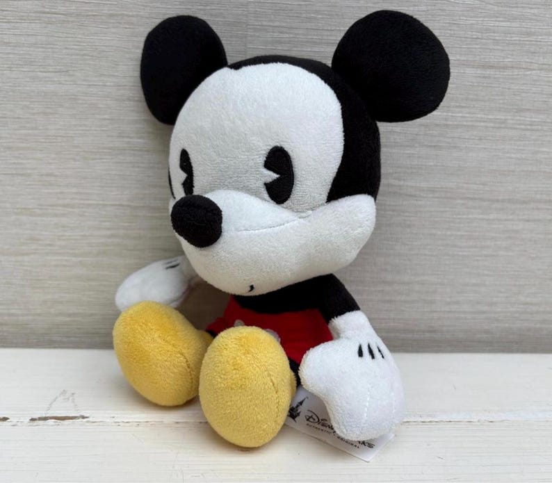 Op de afbeelding: Een pluche Mickey Mouse-speelgoed met zwarte oren, een zwarte neus en een wit gezicht. Het speelgoed heeft een rood shirt, witte handschoenen en gele schoenen. Het speelgoed staat op een wit oppervlak.
