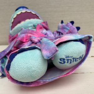 May include: A plush Stitch doll in a galaxy-themed design. The doll features a blue head with a purple mouth, and light blue feet with the word "Stitch" printed on them. The doll is wrapped in a pink, purple, and blue tie-dye fabric.