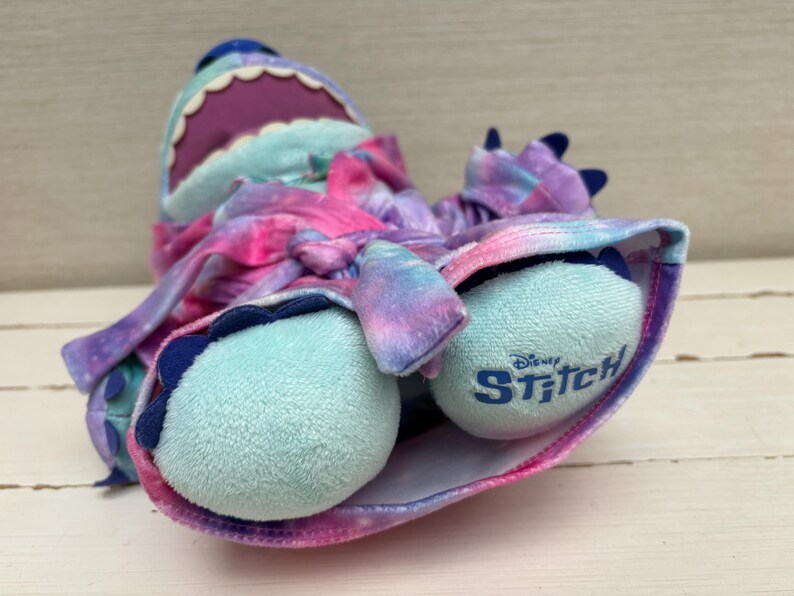 May include: A plush Stitch doll in a galaxy-themed design. The doll features a blue head with a purple mouth, and light blue feet with the word "Stitch" printed on them. The doll is wrapped in a pink, purple, and blue tie-dye fabric.