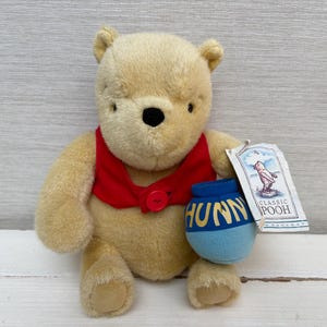 Puede incluir: Un oso de peluche de pelaje color canela claro que lleva un chaleco rojo con un botón. El oso sostiene una pequeña maceta azul con la palabra HUNNY impresa en amarillo. Una etiqueta de papel blanco unida al oso muestra una ilustración y el texto CLASSIC POOH.