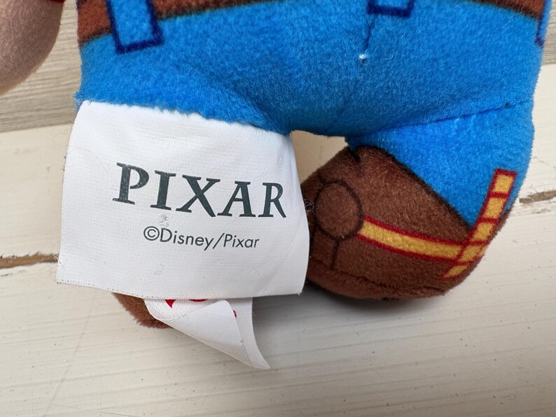 May include: Close-up of a plush toy's leg and tag. The toy has blue pants with brown accents and a brown boot. The tag reads "PIXAR" and "&copy;Disney/Pixar".