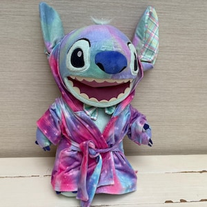May include: A plush Stitch doll wearing a tie-dye robe with a hood. The doll has large ears, a blue nose, and a wide smile. The robe is a mix of pink, purple, and blue colors.