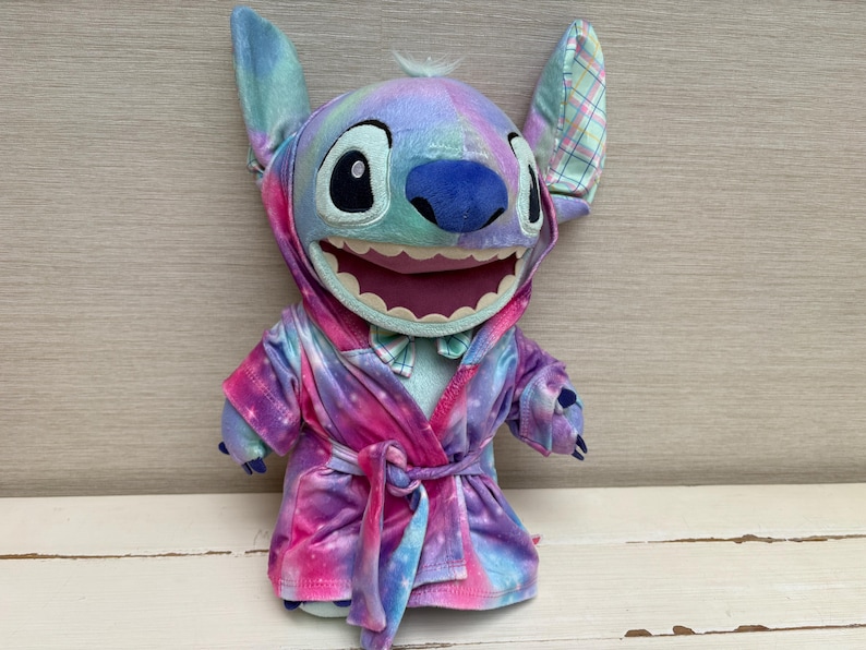 May include: A plush Stitch doll wearing a tie-dye robe with a hood. The doll has large ears, a blue nose, and a wide smile. The robe is a mix of pink, purple, and blue colors.