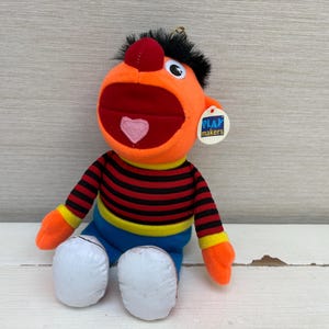 Puede incluir: Una muñeca de peluche con cara naranja, una boca roja grande que contiene una lengua rosa en forma de corazón y cabello negro. La muñeca viste una camisa de manga larga a rayas rojas y negras con puños amarillos, pantalones azules y zapatos blancos. Tiene una etiqueta Playmakers.