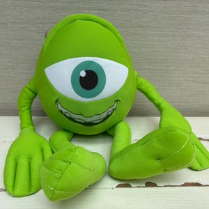 May include: A bright lime green plush toy shaped like a round creature with a single large white eye, a dark teal iris, and a wide smiling mouth showing teeth. The creature has two thin arms with four-fingered hands and two legs with feet.