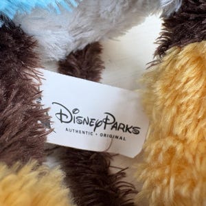 May include: Close-up of a plush toy with brown, yellow, white, and blue fur. A white tag with the Disney Parks logo and the words "AUTHENTIC &bull; ORIGINAL" is visible.