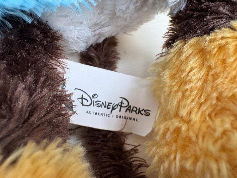 May include: Close-up of a plush toy with brown, yellow, white, and blue fur. A white tag with the Disney Parks logo and the words "AUTHENTIC &bull; ORIGINAL" is visible.