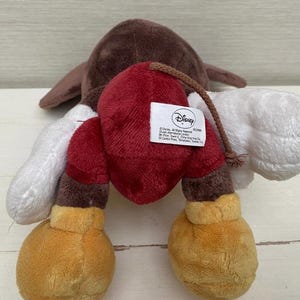 May include: A soft plush toy with a brown head, white arms, and red and brown boxing gloves. The toy has yellow feet and a Disney tag. The toy is made of soft, plush material.