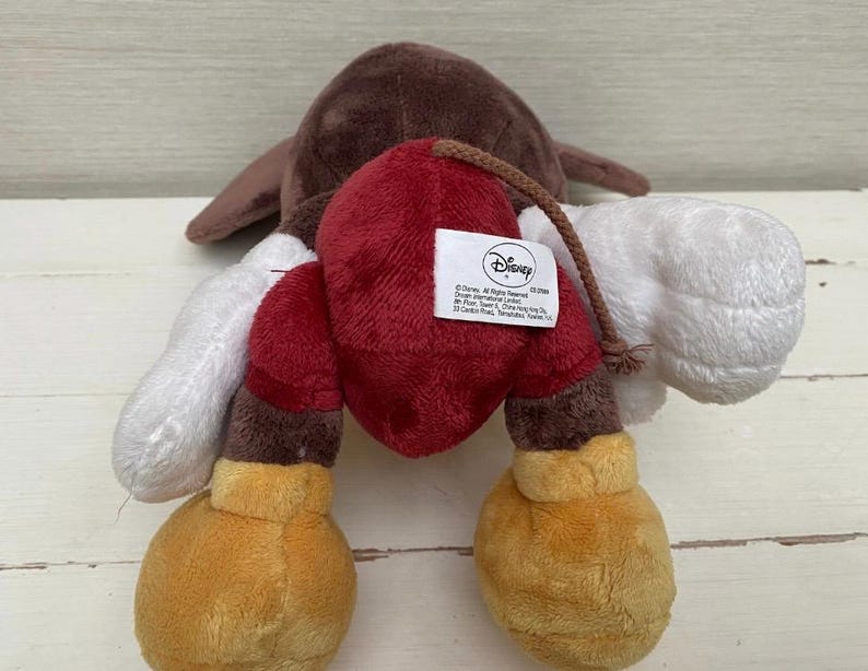 May include: A soft plush toy with a brown head, white arms, and red and brown boxing gloves. The toy has yellow feet and a Disney tag. The toy is made of soft, plush material.