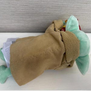 Pode incluir: Um brinquedo de pel&uacute;cia de um personagem de uma popular s&eacute;rie de fic&ccedil;&atilde;o cient&iacute;fica. O brinquedo &eacute; verde claro com orelhas grandes e est&aacute; usando um casaco bege com acabamento marrom. O brinquedo est&aacute; deitado de lado, com uma etiqueta anexada.