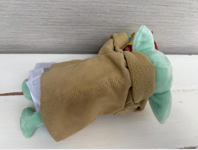 Pode incluir: Um brinquedo de pel&uacute;cia de um personagem de uma popular s&eacute;rie de fic&ccedil;&atilde;o cient&iacute;fica. O brinquedo &eacute; verde claro com orelhas grandes e est&aacute; usando um casaco bege com acabamento marrom. O brinquedo est&aacute; deitado de lado, com uma etiqueta anexada.