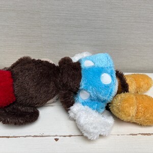 May include: A plush toy in the shape of a cartoon character, featuring brown, red, blue, white, and yellow colors. The toy has a fluffy texture and is lying down. The design includes a red tail, blue body with white spots, and yellow feet.