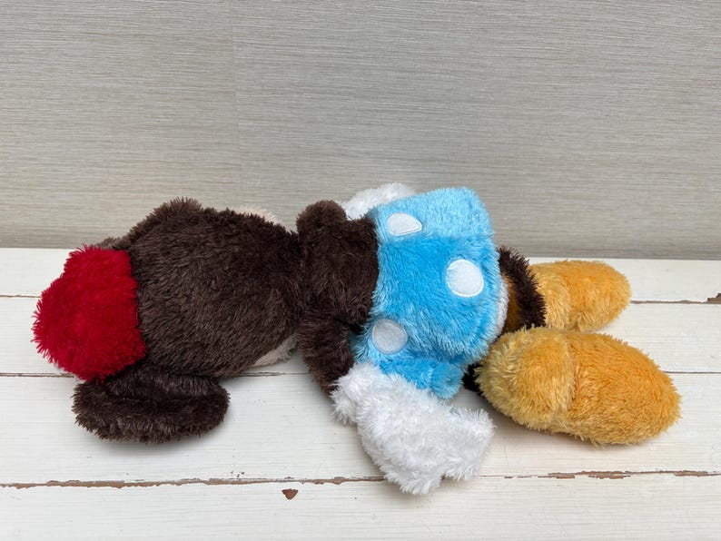 May include: A plush toy in the shape of a cartoon character, featuring brown, red, blue, white, and yellow colors. The toy has a fluffy texture and is lying down. The design includes a red tail, blue body with white spots, and yellow feet.