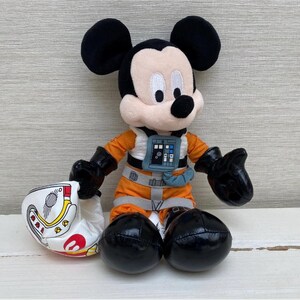 May include: A plush Mickey Mouse toy wearing an orange flight suit and black gloves. The toy is holding a white helmet with red and yellow details. Mickey has black ears and a black nose.