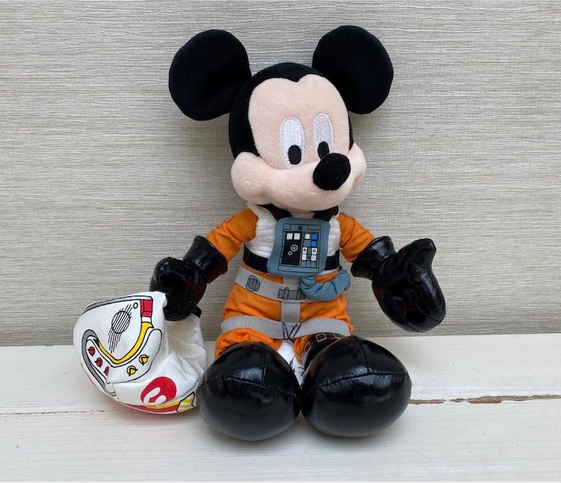 May include: A plush Mickey Mouse toy wearing an orange flight suit and black gloves. The toy is holding a white helmet with red and yellow details. Mickey has black ears and a black nose.