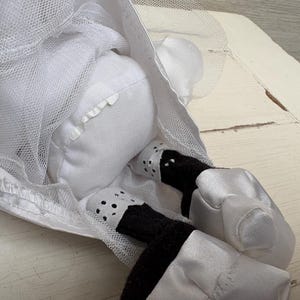 May include: A white stuffed doll wearing a white tulle dress. The doll's legs are black and white, with white satin shoes. The doll is lying down, with its head and upper body visible.