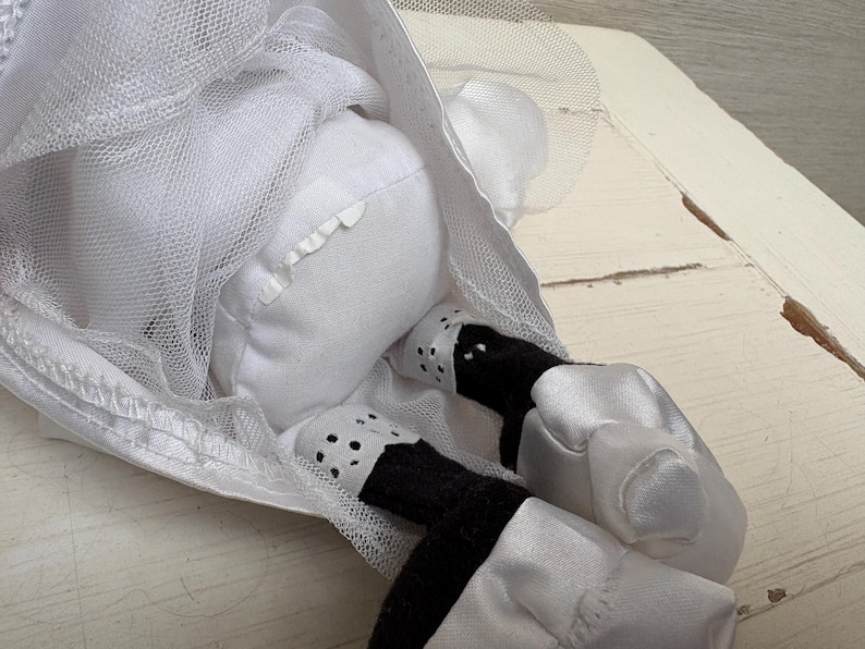 May include: A white stuffed doll wearing a white tulle dress. The doll's legs are black and white, with white satin shoes. The doll is lying down, with its head and upper body visible.