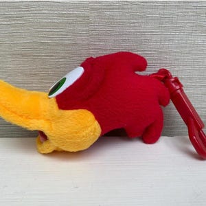May include: A plush Woody Woodpecker head with a yellow beak, red head, and white and green eye. A red plastic clip is attached. The cartoon character is a popular collectible.