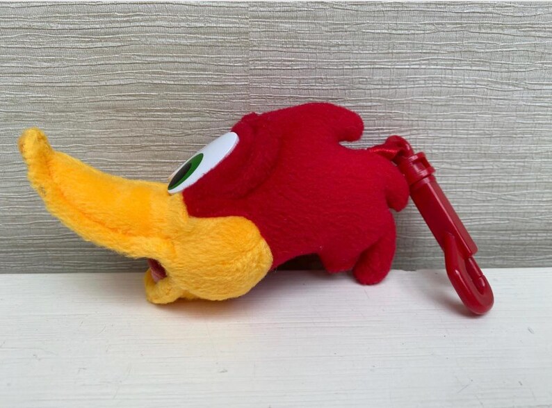 May include: A plush Woody Woodpecker head with a yellow beak, red head, and white and green eye. A red plastic clip is attached. The cartoon character is a popular collectible.