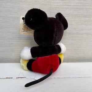 Puede incluir: Un peluche de Mickey Mouse, con cabeza y orejas negras, guantes blancos, pantalones cortos rojos y zapatos amarillos. El juguete tiene una cola negra y una etiqueta adjunta. El juguete est&aacute; sobre una superficie de madera blanca.