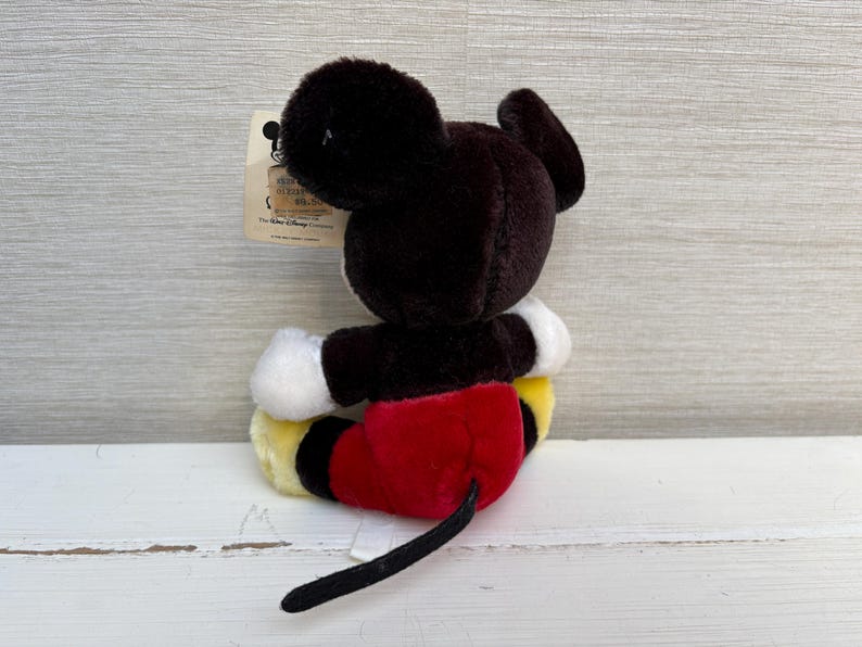 Puede incluir: Un peluche de Mickey Mouse, con cabeza y orejas negras, guantes blancos, pantalones cortos rojos y zapatos amarillos. El juguete tiene una cola negra y una etiqueta adjunta. El juguete est&aacute; sobre una superficie de madera blanca.