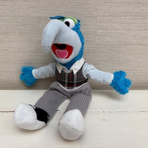 Puede incluir: Un peluche del personaje Gonzo con pelaje azul, una nariz grande y curva y ojos blancos. La figura viste una camisa de manga larga azul claro, un chaleco de cuadros gris oscuro y pantalones de pana gris con zapatos blancos y negros.