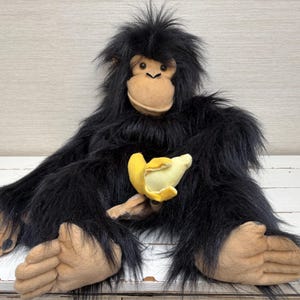 Peut inclure: Une peluche chimpanzé avec une longue fourrure synthétique noire, un visage, des mains et des pieds beiges, tenant une petite banane jaune et crème. Le chimpanzé a des yeux sombres et une apparence douce, assis en position droite.