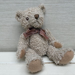 May include: A soft teddy bear with light brown fur and a brown bow. The bear has a textured appearance, with tan-coloured paws and a matching snout. The bear is sitting on a white wooden surface.