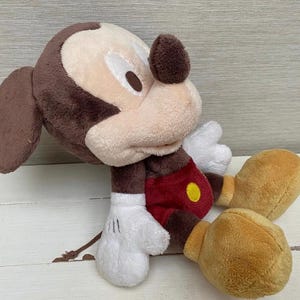 May include: A plush Mickey Mouse toy in a seated position. The toy has a brown head and ears, a tan face, white gloves, red shorts with a yellow button, and tan shoes. The toy is made of soft, plush material.