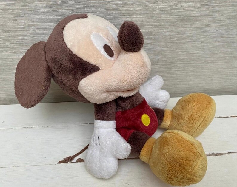 May include: A plush Mickey Mouse toy in a seated position. The toy has a brown head and ears, a tan face, white gloves, red shorts with a yellow button, and tan shoes. The toy is made of soft, plush material.