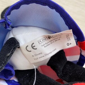 Puede incluir: Primer plano de un peluche con un sombrero azul y rojo con un dise&ntilde;o de la bandera brit&aacute;nica. El juguete tiene orejas negras y una nariz negra. Una etiqueta blanca con el logotipo de Disney es visible.
