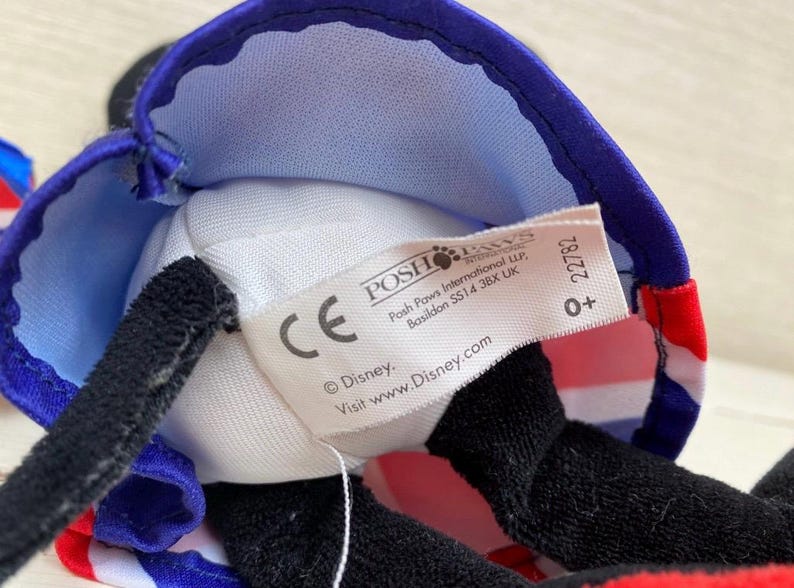 Puede incluir: Primer plano de un peluche con un sombrero azul y rojo con un dise&ntilde;o de la bandera brit&aacute;nica. El juguete tiene orejas negras y una nariz negra. Una etiqueta blanca con el logotipo de Disney es visible.