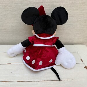 May include: A plush Minnie Mouse doll, seen from the rear, featuring black ears, a red bow, and a red dress adorned with white polka dots. The doll has white gloves and a black tail. The background is a light-coloured wooden surface.