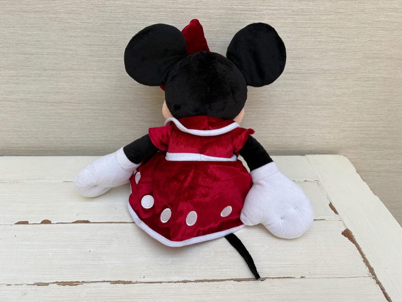May include: A plush Minnie Mouse doll, seen from the rear, featuring black ears, a red bow, and a red dress adorned with white polka dots. The doll has white gloves and a black tail. The background is a light-coloured wooden surface.