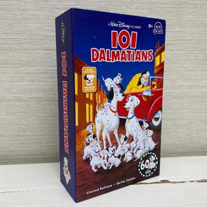 Può includere: Un cofanetto del classico Disney "La carica dei 101". La scatola presenta uno sfondo blu scuro con il titolo in rosso e bianco. L'immagine mostra una scena con cani dalmata e Crudelia De Mon in un'auto rossa. Include un peluche.